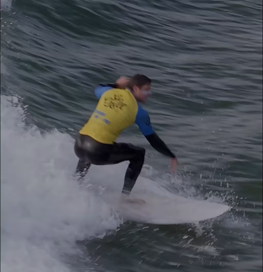 A man in a black wetsuit and a blue and yellow vest surfing the waves in Carcavelos beach on a white surf board.